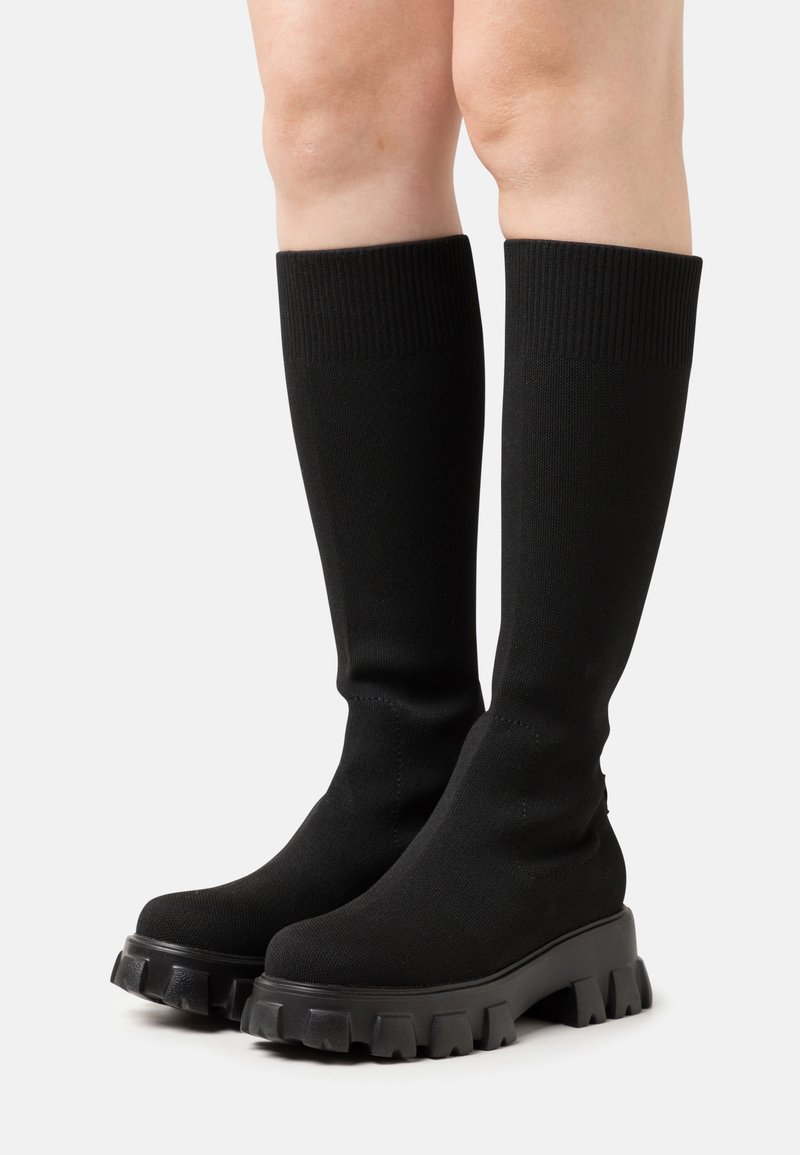 Botas negras hasta la rodilla hechas de un tejido elástico, con punta redondeada y suela de goma gruesa con dibujos pronunciados.