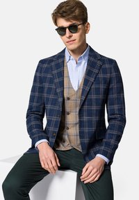 Granatowa marynarka w kratę z brązowymi akcentami, nałożona na beżową kamizelkę, noszona z jasnoniebieską koszulą. Ciemnozielone spodnie dopełniają outfit.