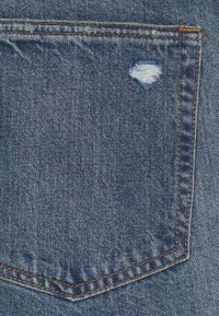 Gros plan sur la poche arrière d’un jean en denim bleu avec des coutures orange et un petit trou usé près du coin supérieur droit.