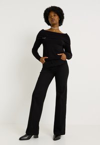 Mujer con peinado afro que lleva una blusa negra off-shoulder, pantalones anchos negros, botas negras y un collar de plata, de pie contra un fondo liso.