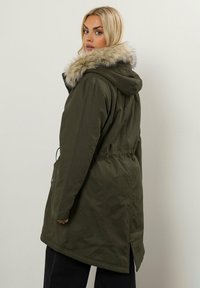 Olivgrön parkajacka med en fuskpälsfodrad huva, insydd midja och sidofickor. Har en lång, rak skärning och bakre ventil.