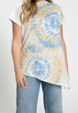 Femme portant un haut asymétrique à manches courtes tie-dye bleu et jaune avec un jean bleu clair sur fond uni.
