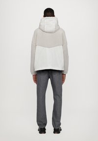 Giacca in fleece grigia con cappuccio, presenta un pannello di tessuto più leggero nella parte inferiore, abbinata a pantaloni in denim grigi e scarpe nere.