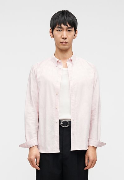 Ροζ ανοιχτό πουκάμισο με ρίγες και κουμπιά, μακριά μανίκια, συνοδευόμενο από λευκή φανέλα και μαύρο παντελόνι, με ασημένια αγκράφα στη ζώνη.