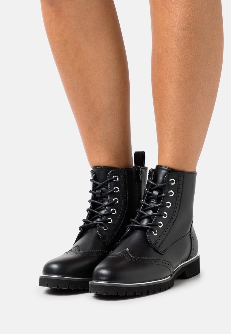 Bottes basses en cuir noir à lacets avec détails brogue et fermeture éclair latérale portées sur des jambes nues sur fond blanc.