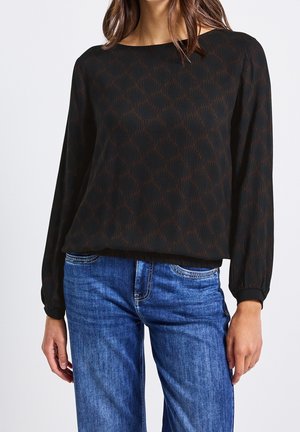 Vrouw draagt een zwart bloesje met lange mouwen met subtiel geometrisch patroon en blauwe spijkerbroek, staand tegen een effen achtergrond.