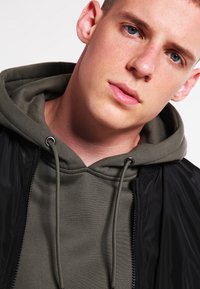 Zwarte bomberjack met een rits aan de voorkant, gedragen over een olijfgroene hoodie. De hoodie heeft verstelbare trekkoorden en een soepele textuur.