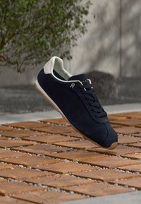 Δερμάτινα αθλητικά παπούτσια σε ναυτικό μπλε με άσπρο τακούνι, με μαύρα κορδόνια, χρυσό λογότυπο και λαστιχένια σόλα σε ξύλινη επιφάνεια.