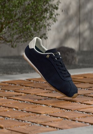 Baskets en daim bleu marine avec un talon blanc, dotées de lacets noirs, d'un accent logo doré et d'une semelle en caoutchouc sur une surface en bois.