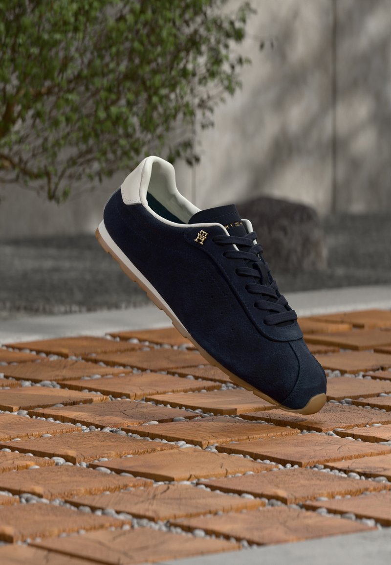 Δερμάτινα αθλητικά παπούτσια σε ναυτικό μπλε με άσπρο τακούνι, με μαύρα κορδόνια, χρυσό λογότυπο και λαστιχένια σόλα σε ξύλινη επιφάνεια.