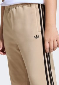 Beige tracksuit pants with black side stripes, elastic waistband, and a small black Adidas logo on the front pocket area. Soft material.