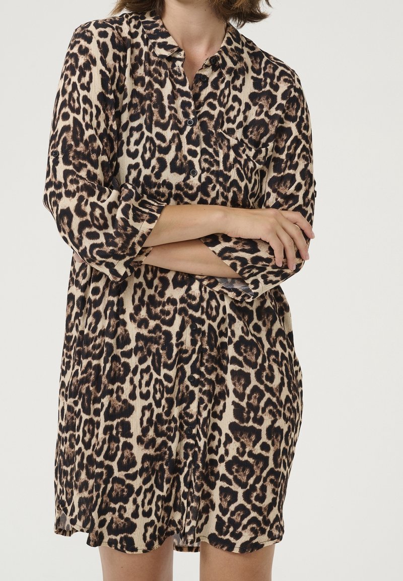Femme portant une robe boutonnée à manches longues avec un imprimé léopard marron et noir, bras croisés, sur un fond clair uni.