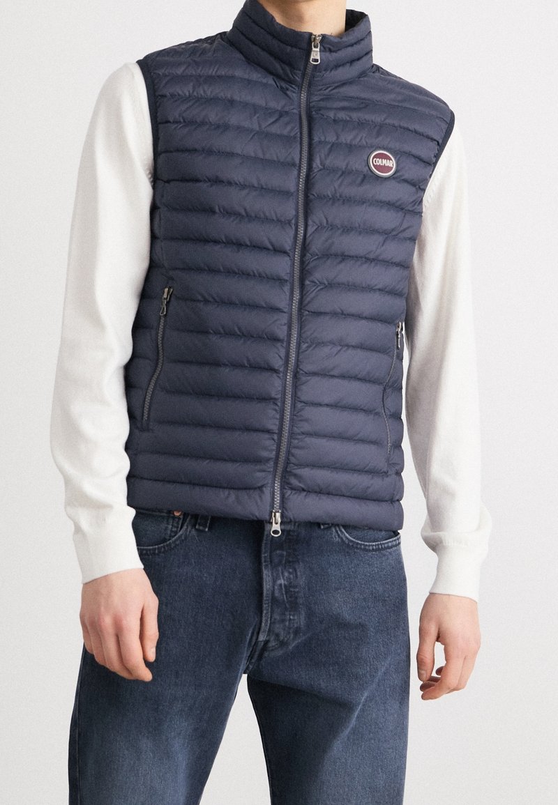 Navy puffer vest with vertical quilted pattern, high collar, front zipper, and two side pockets. Worn over a white long-sleeve shirt.
