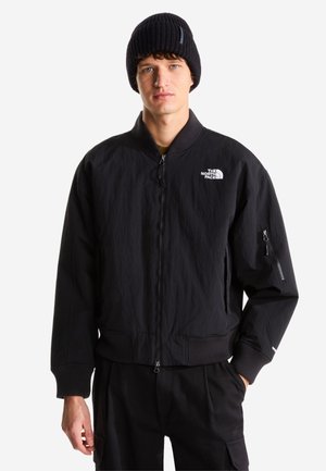 Jeune homme portant un bonnet noir et une veste bomber noire North Face avec une poche zippée sur la manche, sur fond blanc.