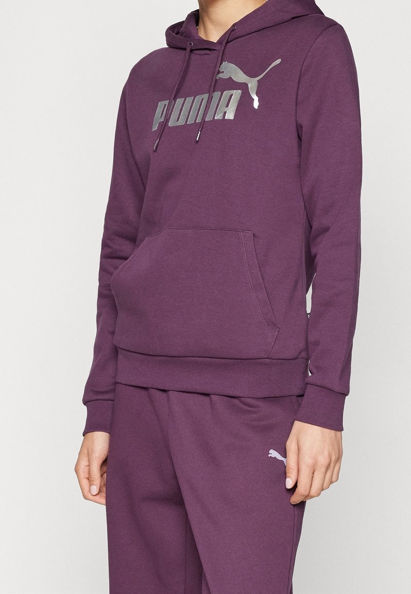 Personne portant un sweat à capuche violet Puma avec une poche avant et un pantalon violet assorti avec le logo Puma sur la cuisse.