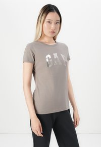 Young woman with long ombre hair wearing a taupe GAP t-shirt and black pants, standing against a plain white background.