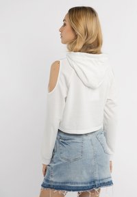 Felpa bianca cropped con spalle scoperte e cappuccio, abbinata a una gonna di jeans sfilacciata. Il tessuto appare morbido e strutturato.