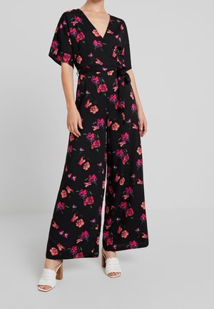 Kvinde iført sort buksedragt med vide ben og blomsterprint, korte ærmer, rund halsudskæring, bånd i taljen og hvide hælede sandaler mod en ensfarvet baggrund.