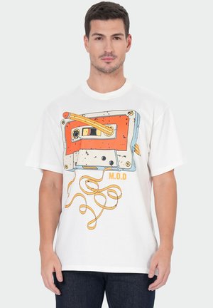 Man draagt een wit T-shirt met een grote afbeelding van een ontwarde oranje en gele cassetteband en de letters "M.O.D" aan de voorkant.