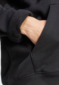 Sweat à capuche noir avec des poignets texturés, une poche avant, et un tissu doux et lisse. Les coutures sont visibles le long des seams.