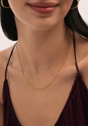 Mujer con un collar de cadena delgada de oro, pendientes de aro dorados y un top de tirantes finos de color púrpura oscuro, con piel suave visible.
