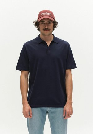 Muž s kučeravými vlasmi a fúzmi, ktorý nosí červenú čiapku s nápisom "Noosa Beach", tmavomodré polo tričko a svetlomodré džínsy na bielom pozadí.