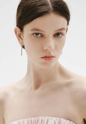 Joven con ojos azules y cabello oscuro, que lleva pendientes de plata colgantes y un top sin tirantes de color rosa pálido, sobre un fondo blanco.