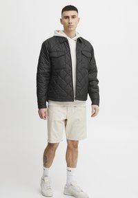 Svart quiltet jakke med diamantmønster, frontglidelås, to brystlommer, lagd over en lys farget hettegenser og paret med beigefargede shorts.