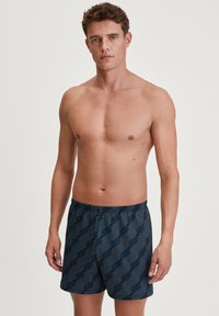 Patronen zwembroek in donkerblauw, met geometrische vormen in lichter blauw. Elastische tailleband, losse pasvorm, lengte tot halverwege het dijbeen.