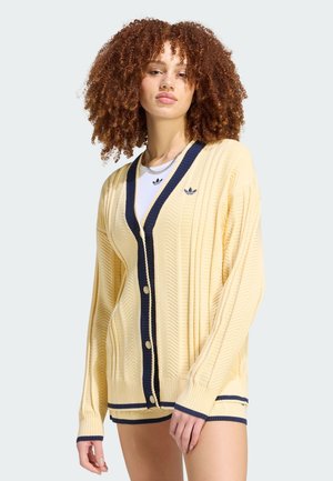 Mujer con cabello rizado que lleva un cárdigan amarillo abotonado con ribete azul marino sobre una camisa blanca, de pie contra un fondo liso.
