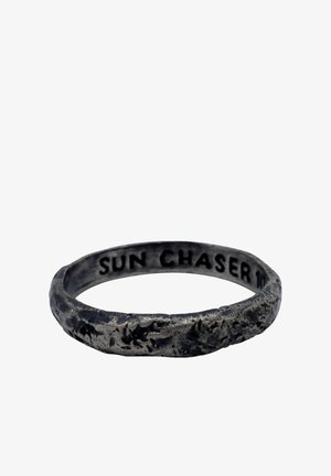 Metallinen sormus, jossa on teksturoitu, mattamusta viimeistely. Sisäpuolella kaiverrettu teksti "SUN CHASER." Yksinkertainen, pyöreä muoto epätasaisella pinnalla.