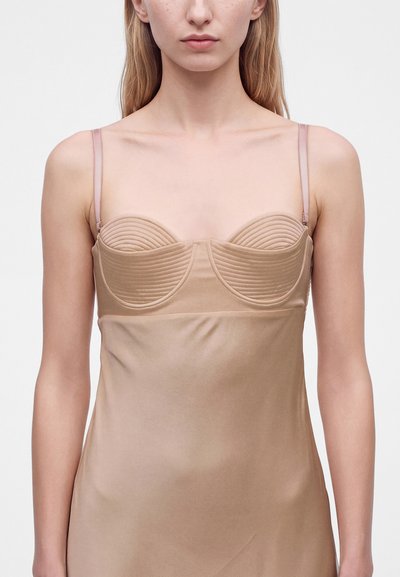 Femme portant une robe nuisette en satin beige avec de fines bretelles et des bonnets arrondis et matelassés, montrée des épaules jusqu'au milieu du torse.