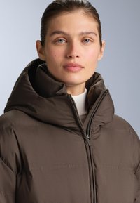 Hnedá nafukovacia bunda s vysokou kapucňou, viditeľným zipsom a hladkou textúrou. Látka má jemný lesk a je mierne polstrovaná.