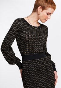 Svart stickad klänning med guldiga zigzag-mönster, rundad halsringning och puffärmar. Ribbad midja ger struktur åt designen.