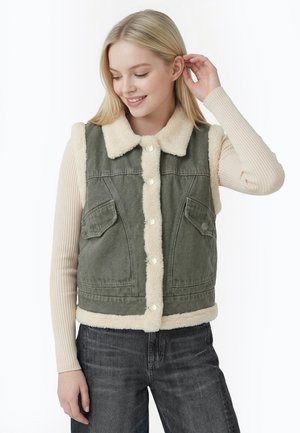 Ung kvinde iført en grøn knappet vest med hvid pelskant over en ribbet beige langærmet top og mørke jeans, rører ved sit hår.