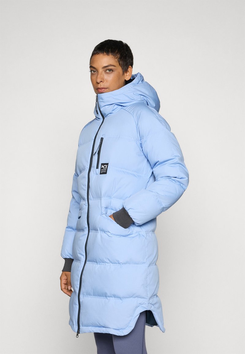 Cappotto imbottito blu con cappuccio, dotato di zip completa, tasche frontali e un logo sul petto. Le maniche si estendono con un accento grigio.