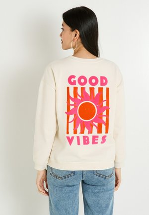 Femme aux longs cheveux noirs portant un sweat crème avec un graphique de soleil rose et orange et le texte "GOOD VIBES" au dos, un jean bleu et des boucles d'oreilles créoles.
