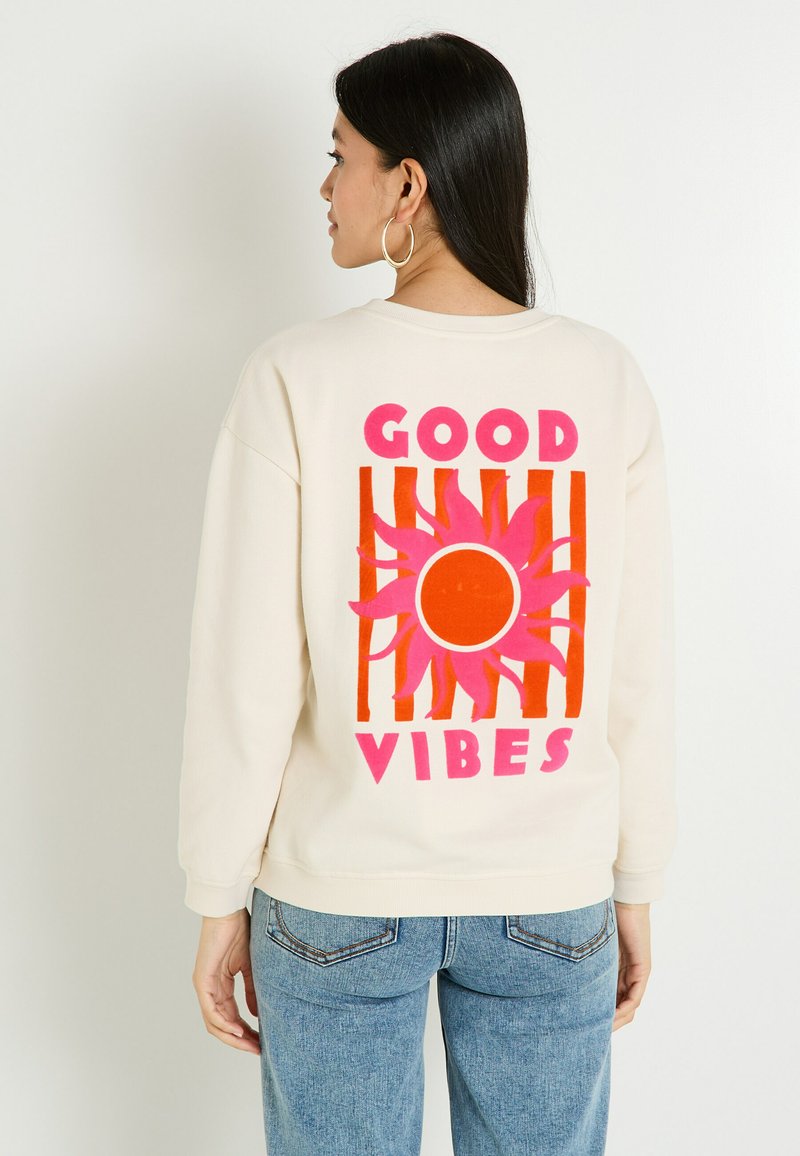 Femme aux longs cheveux noirs portant un sweat crème avec un graphique de soleil rose et orange et le texte "GOOD VIBES" au dos, un jean bleu et des boucles d'oreilles créoles.