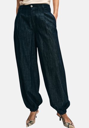 Donna con pantaloni di denim scuro e larghi con polsini raggruppati e scarpe con tacco alto con stampa a motivo serpente, in piedi con le mani in tasca.