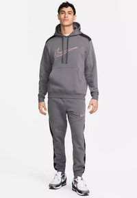 Felpa grigia Nike con logo rosa e accenti neri, abbinata a pantaloni della tuta grigi con strisce laterali nere. Tessuto a rete, vestibilità atletica.