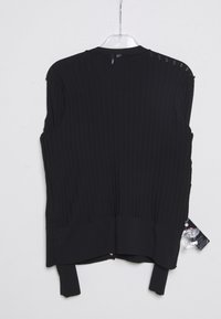 Pull sans manches noir en maille avec ourlet côtelé suspendu à un cintre en plastique transparent contre un mur blanc, avec une étiquette de vêtement attachée sur le côté.