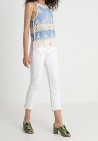 Ljusblå ärmlös blus med beige spetsdetaljer, kombinerad med vita jeans och olivgröna klackskor.