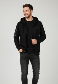 Schwarze Zip-Hoodie mit Kordelzügen, dezentes Textdesign am linken Arm, kombiniert mit einem schwarzen T-Shirt und dunklen Jeans. Glatter Stofftextur.