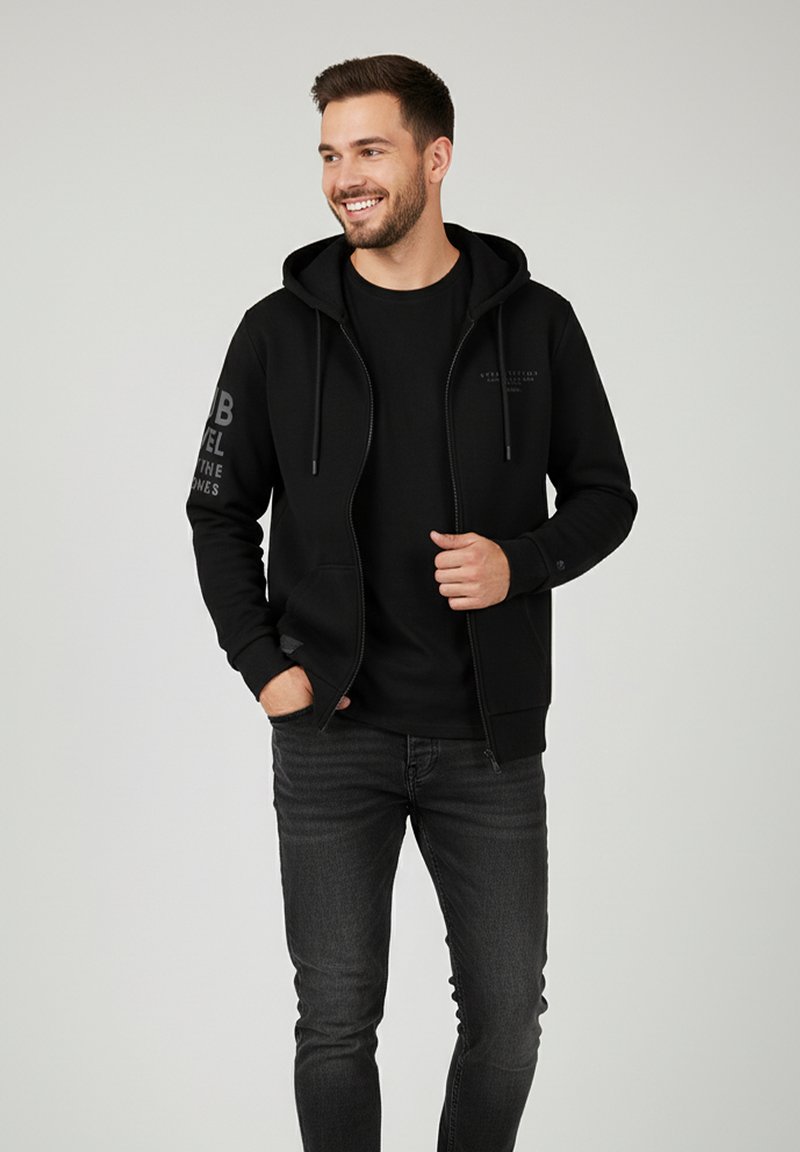 Schwarze Zip-Hoodie mit Kordelzügen, dezentes Textdesign am linken Arm, kombiniert mit einem schwarzen T-Shirt und dunklen Jeans. Glatter Stofftextur.