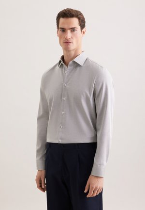 Mand stående iført lysegrå skjorte med knapper og lange ærmer samt mørkeblå bukser, neutral baggrund.