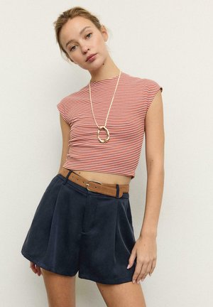 Donna che indossa un top corto senza maniche a righe rosse e bianche, pantaloncini navy a vita alta con una cintura marrone e una collana con ciondolo dorato.