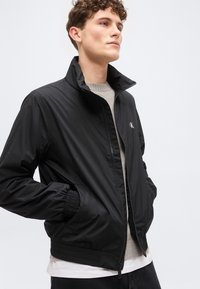 Νέος άνδρας με σγουρά μαλλιά που φοράει μαύρο μπουφάν με φερμουάρ πάνω από μπεζ πουλόβερ, με τα χέρια στις τσέπες, μπροστά σε απλό φωτεινό φόντο.