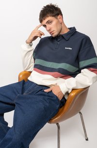 Marineblauwe en crèmekleurige polo met kleurgeblokte groene en bordeauxrode strepen, gemaakt van zachte stof. Model zit in een houten stoel met de hand op het voorhoofd.