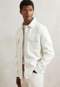 Chaqueta de forro polar blanca con botones a presión, que cuenta con dos bolsillos de pecho de pana. Textura suave y corte relajado, llevada sobre una blusa ligera.