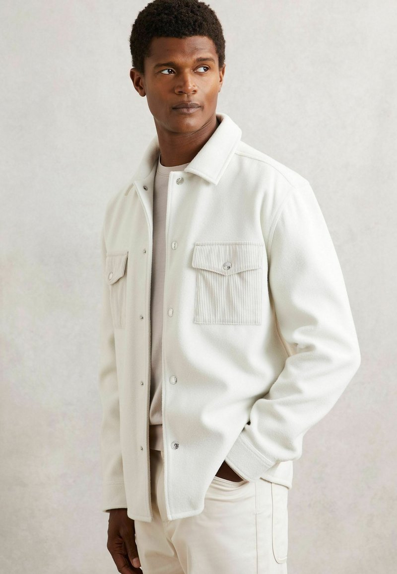 Chaqueta de forro polar blanca con botones a presión, que cuenta con dos bolsillos de pecho de pana. Textura suave y corte relajado, llevada sobre una blusa ligera.